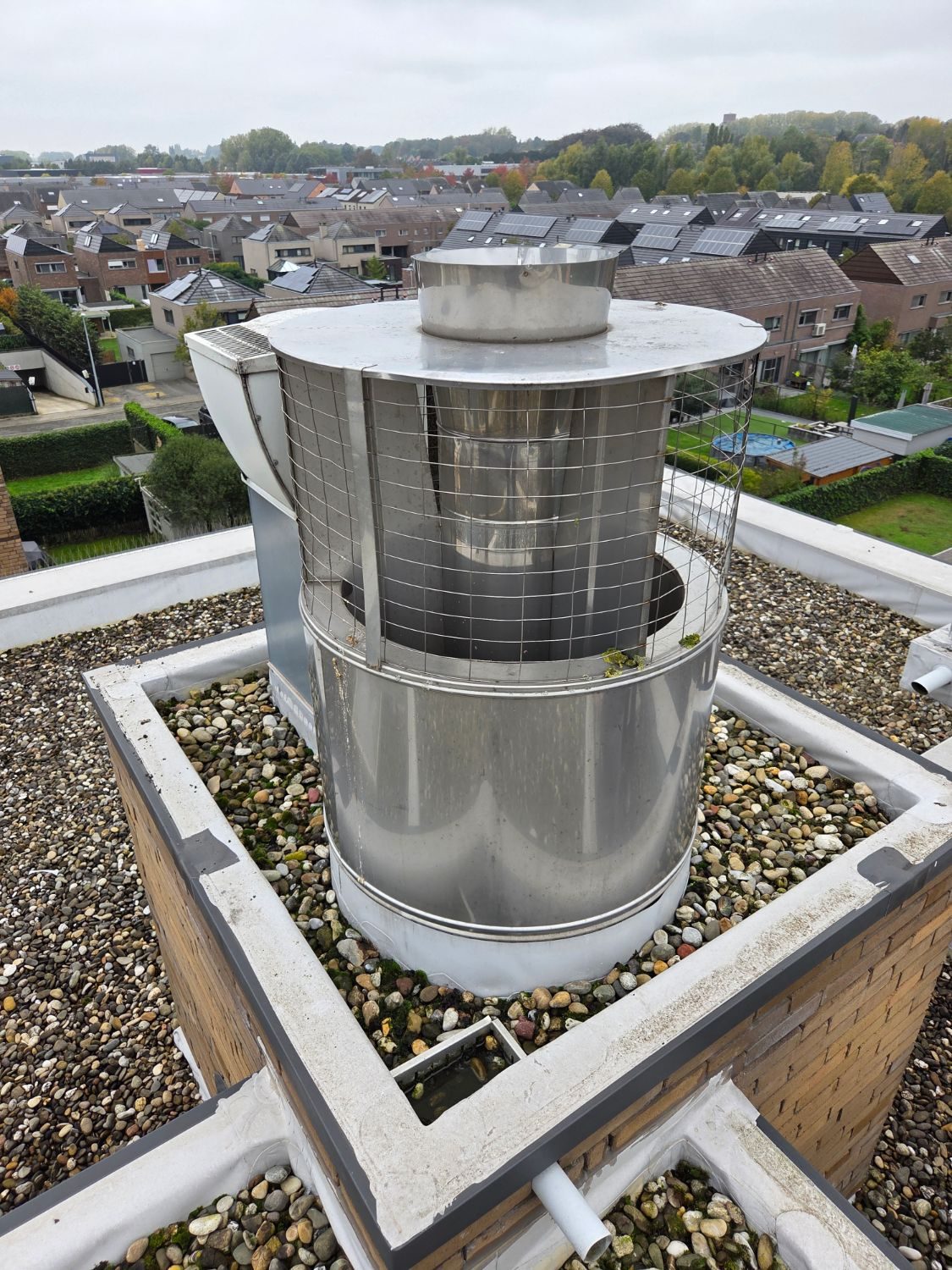 Oude CLV-kap op het platte dak van een appartementsgebouw: inox trommel met een band gaas, plat deksel en de rookgasbuis in het midden